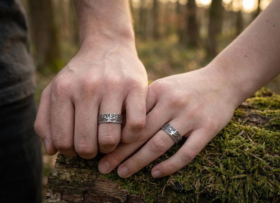 A clean studio shot of two matching sterling silver rings resting on a slate grey surface. Both bands feature a continuous panoramic design of a tree with spreading branches and deep roots. The tree motif is polished silver, which contrasts sharply with the textured, oxidized grey background that resembles tree bark or drifting soil.