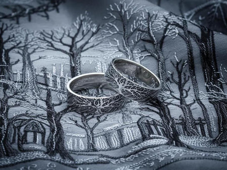 A clean studio shot of two matching sterling silver rings resting on a slate grey surface. Both bands feature a continuous panoramic design of a tree with spreading branches and deep roots. The tree motif is polished silver, which contrasts sharply with the textured, oxidized grey background that resembles tree bark or drifting soil.