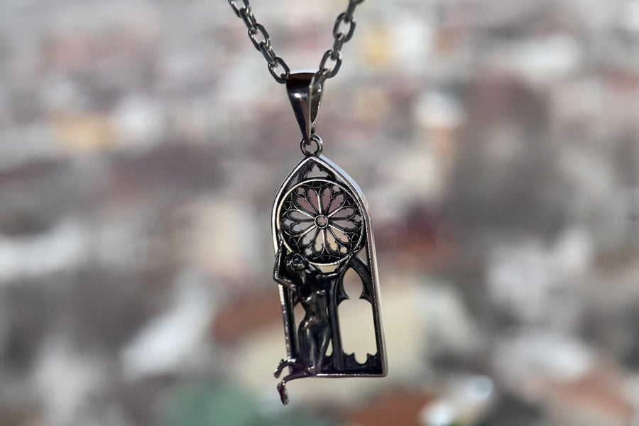 A cinematic studio shot of a tall, arched sterling silver pendant. The frame mimics a Gothic cathedral window. Inside the arch, a sculpted female figure (appearing nude) kneels or floats, reaching up to hold a large, circular "Rose Window" tracery wheel above her head. The silver is heavily oxidized, creating deep shadows behind the figure and the window spokes. (etsy image id: 7448666740)
