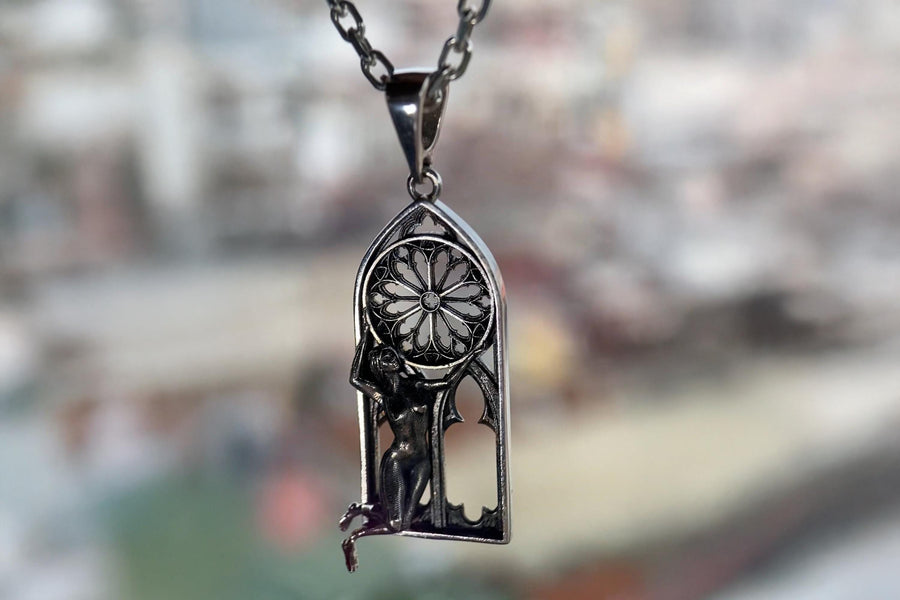 A cinematic studio shot of a tall, arched sterling silver pendant. The frame mimics a Gothic cathedral window. Inside the arch, a sculpted female figure (appearing nude) kneels or floats, reaching up to hold a large, circular "Rose Window" tracery wheel above her head. The silver is heavily oxidized, creating deep shadows behind the figure and the window spokes. (etsy image id: 7448666740)