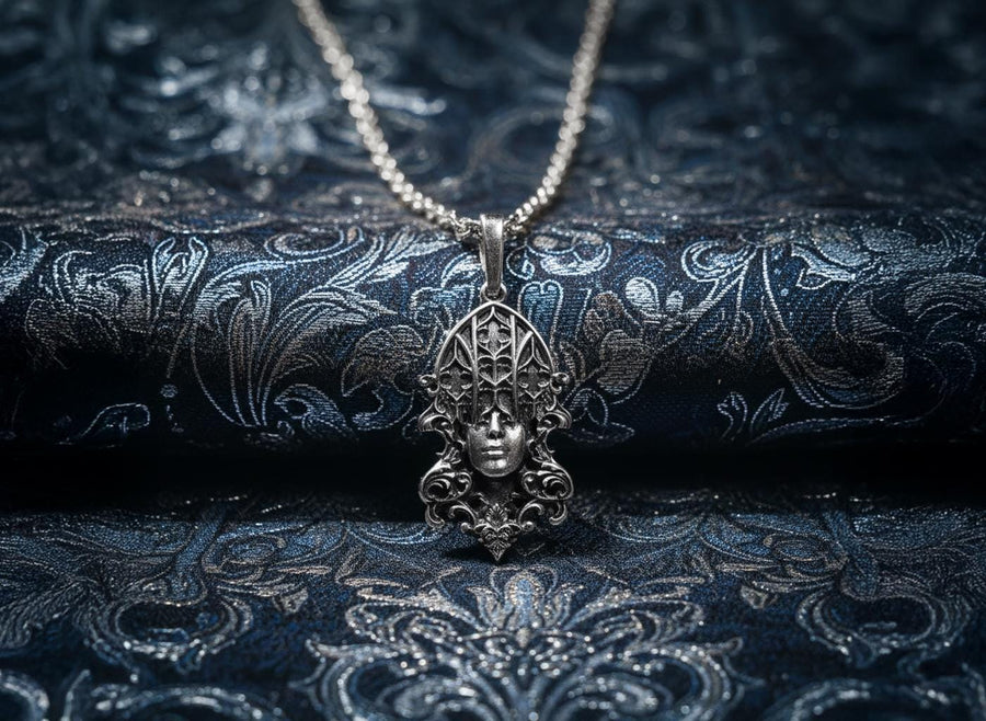 A detailed studio shot of a sterling silver pendant resting on a dark, textured stone surface. The design features a realistic human face in the center, but the forehead and eyes are obscured by a grid of Gothic window tracery (vertical bars and trefoils). The chin is framed by swirling Art Nouveau-style acanthus leaves. The silver is heavily oxidized, giving the face the appearance of a weathered stone statue. (etsy image id: 7448678222)