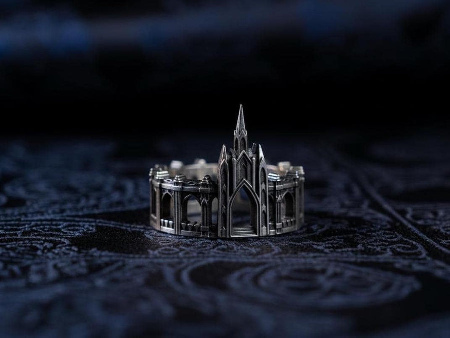 A cinematic studio shot of a sculptural sterling silver ring standing on a reflective surface. The ring is shaped like a miniature building, featuring a tall central spire that rises to a sharp point. The band itself is formed by a colonnade of pointed Gothic arches and buttresses that wrap around the finger. The silver is heavily oxidized, creating deep shadows in the "windows" and "doorways" of the structure. (etsy image id: 7450125356)