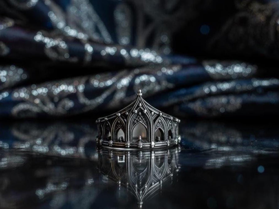 A cinematic studio shot of a sculptural sterling silver ring standing on a reflective surface. The ring is designed as a "crown" or "coronet," formed by a continuous circle of Gothic cathedral arches. The central arch is the tallest, creating a focal point like a spire. The silver is heavily oxidized, with black recesses emphasizing the ribbed vaulting and architectural details against the polished ridges.