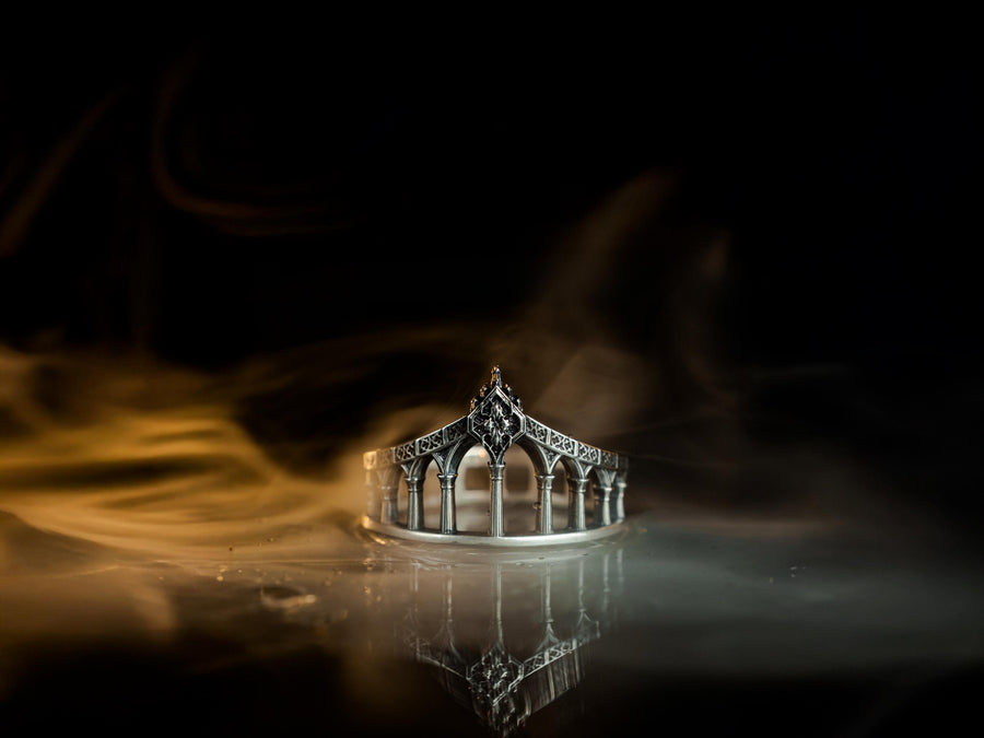 A cinematic studio shot of a sculptural sterling silver ring standing in shallow water. The design mimics a bridge or aqueduct, featuring a row of vertical pillars or columns that support a rising upper rail. The center rises to a sharp point adorned with a floral or quatrefoil relief. The space between the columns is open, creating a "see-through" architectural silhouette. (etsy image id: 7450247674)