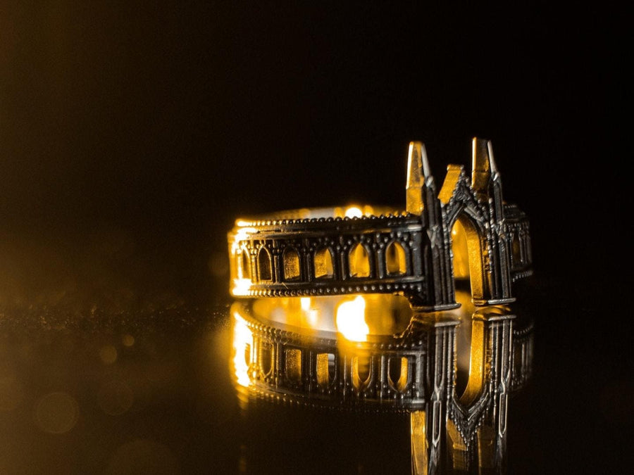 A cinematic studio shot of a sculptural sterling silver ring standing on a reflective surface. The design mimics the facade of a Gothic cathedral or castle gate. Two tall, pointed spires rise up on either side of a central archway. The band continues outwards with a colonnade of smaller arches. The silver features a beaded "cornice" detail along the edges and is heavily oxidized to emphasize the depth of the "stonework." (etsy image id: 7450300962)