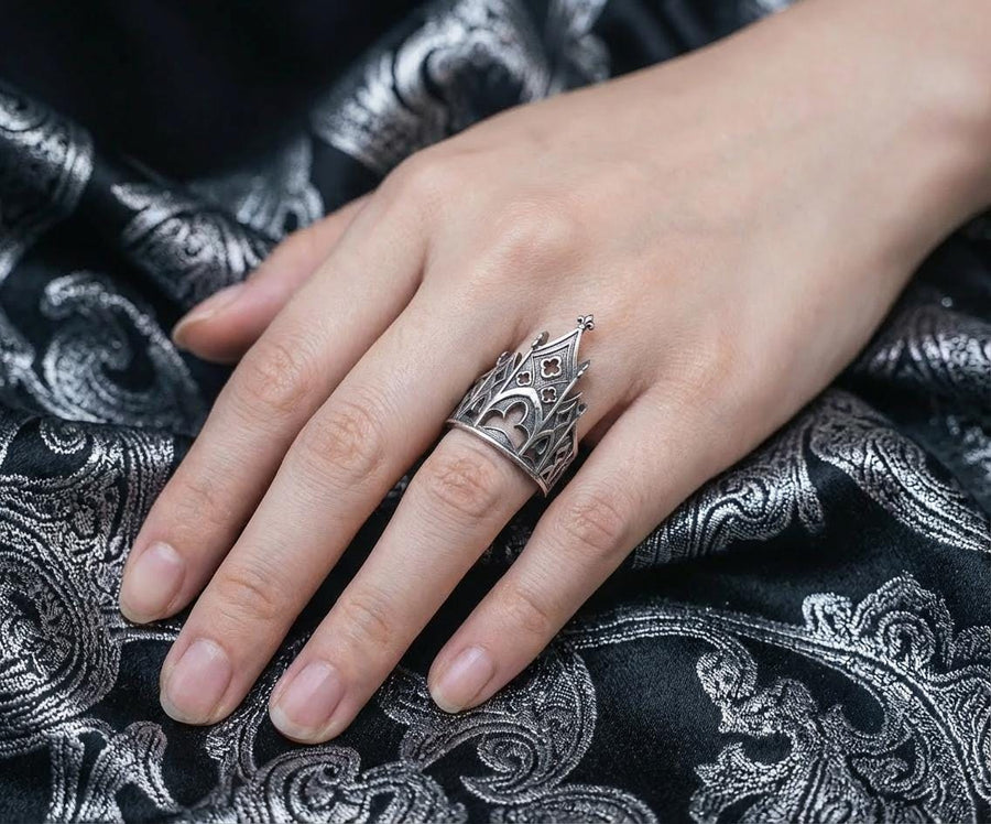 A clean studio shot of a sculptural sterling silver ring resting on a matte grey surface. The ring depicts a Gothic building facade with a tall, triangular central peak (gable) filled with geometric cross-shaped tracery. Flanking the center are lower pointed arches and vertical buttresses that wrap around the band. The silver is matte-finished with dark oxidation in the recesses to mimic stone shadows. (etsy image id: 7452370200)