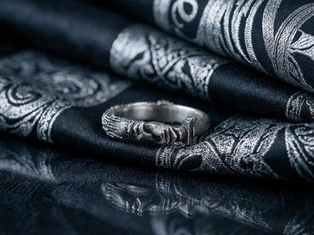 A cinematic studio shot of a sculptural sterling silver ring resting on a dark textured fabric. The focal point is two realistic hands clasped in a handshake (the Fede motif). Instead of fabric sleeves, the wrists emerge from architectural column capitals with distinct "volute" scrolls (Ionic style). The band continues as a textured, fluted column shaft. The silver is heavily oxidized to mimic ancient stone. (etsy image id: 7452415972)