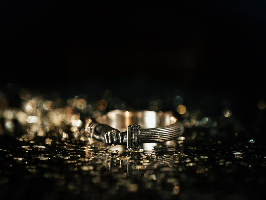 A cinematic studio shot of a sculptural sterling silver ring resting on a dark textured fabric. The focal point is two realistic hands clasped in a handshake (the Fede motif). Instead of fabric sleeves, the wrists emerge from architectural column capitals with distinct "volute" scrolls (Ionic style). The band continues as a textured, fluted column shaft. The silver is heavily oxidized to mimic ancient stone. (etsy image id: 7452415972)