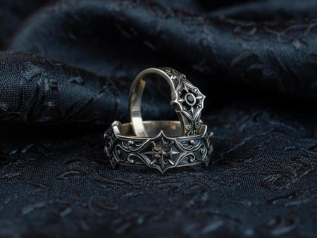 A cinematic studio shot of a matching two-piece ring set on dark velvet. The top ring stands upright, featuring a diamond-shaped face with a four-pointed star design and a small, round black stone in the center. The bottom ring lies flat, displaying a wide band with a repeating pattern of the same star/cross motif connected by swirling filigree. The silver is polished on the ridges and dark in the background.