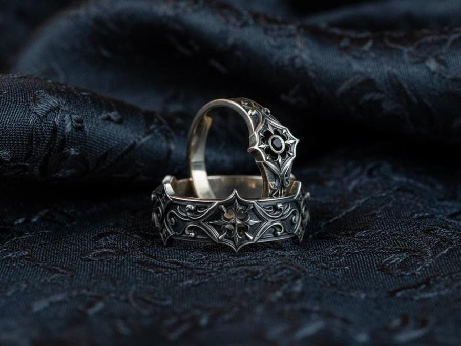 A cinematic studio shot of a matching two-piece ring set on dark velvet. The top ring stands upright, featuring a diamond-shaped face with a four-pointed star design and a small, round black stone in the center. The bottom ring lies flat, displaying a wide band with a repeating pattern of the same star/cross motif connected by swirling filigree. The silver is polished on the ridges and dark in the background.