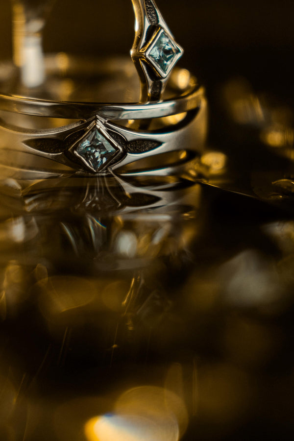 This Art Deco wedding ring set features a sterling silver engagement ring and matching band with bold geometric cutouts and a bright blue square gemstone. The clean lines, layered shapes, and sculptural silhouette create a modern yet vintage feel ideal for couples seeking unique, architecturally inspired bridal jewelry captured on a reflective surface for elegant presentation.