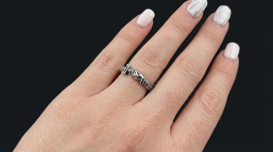 A cinematic studio shot of a sculptural sterling silver ring resting on a dark textured fabric. The focal point is two realistic hands clasped in a handshake (the Fede motif). Instead of fabric sleeves, the wrists emerge from architectural column capitals with distinct "volute" scrolls (Ionic style). The band continues as a textured, fluted column shaft. The silver is heavily oxidized to mimic ancient stone. (etsy image id: 7452415972)