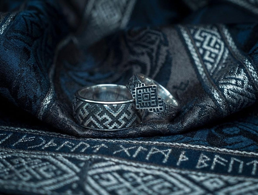 A studio shot of a two-piece sterling silver ring set with a high-contrast black and silver finish. The standing ring features a square, diamond-shaped face created by stepped geometric lines. The lying ring is a wide band with a continuous "zig-zag" or "step" pattern running around the circumference. The design mimics the look of cross-stitch embroidery or pixelated ancient art rendered in metal.