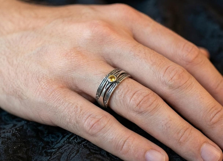 A detailed studio shot of a wide, heavy-gauge sterling silver ring. The focal point is a round, deep red gemstone (Garnet) bezel-set in the center. Flanking the stone are three parallel, tapered grooves that resemble the fletching of an arrow or the futuristic paneling of armor. The metal surfaces are brushed silver, while the deep grooves are blackened with oxidation to emphasize the directional lines. (etsy image id: 7441158070)