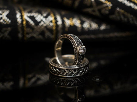 A warm, atmospheric shot of a matching wedding ring set. The engagement ring stands upright, showcasing a round white gemstone in a bezel setting. Lying flat beneath it is a matching wide band without a stone. Both rings share a cohesive design: a central diagonal "basket weave" pattern flanked by twisted rope borders. The silver is oxidized to reveal the intricate texture of the weave.
