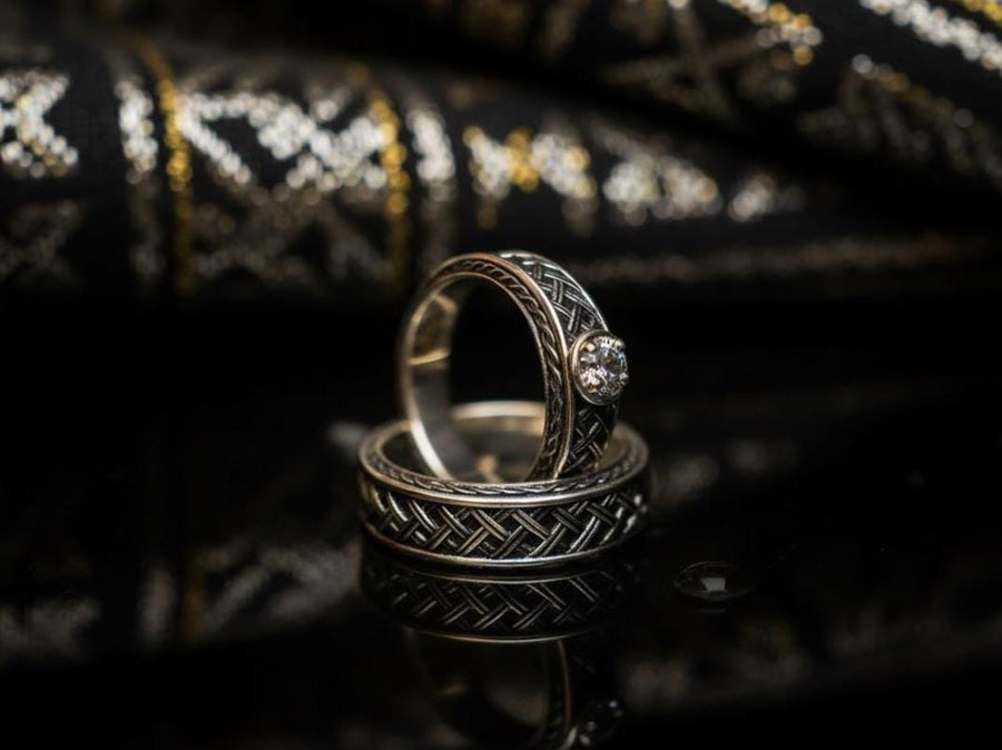 A warm, atmospheric shot of a matching wedding ring set. The engagement ring stands upright, showcasing a round white gemstone in a bezel setting. Lying flat beneath it is a matching wide band without a stone. Both rings share a cohesive design: a central diagonal "basket weave" pattern flanked by twisted rope borders. The silver is oxidized to reveal the intricate texture of the weave.