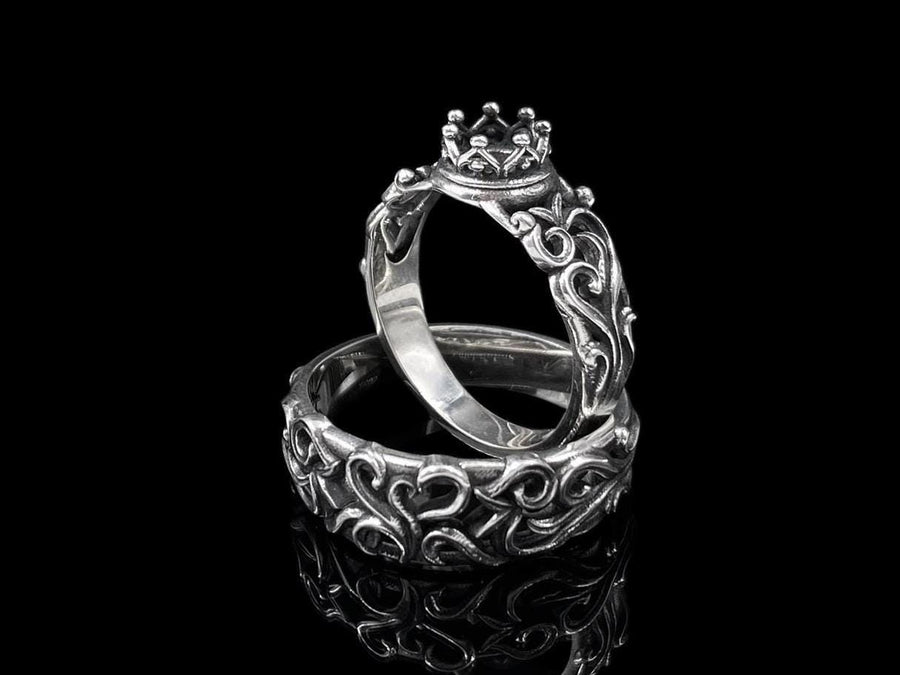 A high-contrast studio shot of a two-piece sterling silver ring set. The top ring features a raised, three-dimensional "crown" or "coronet" motif with small spheres on the tips. The bottom ring is a matching wide band. Both rings are covered in intricate, raised scrollwork or vine-like filigree. The silver is polished bright on the raised swirls, contrasting with the black oxidized background.