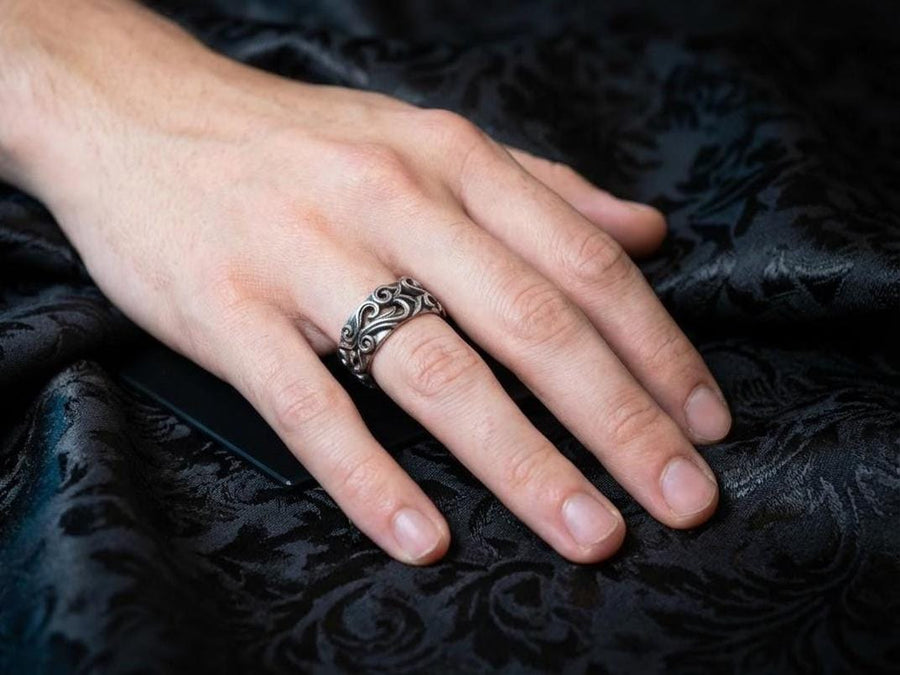 A detailed close-up of a wide sterling silver ring resting on a reflective surface. The band is covered in a deep-relief filigree pattern consisting of interlocking scrolls, vines, and subtle heart shapes. The raised designs are polished bright silver, while the recessed background is heavily oxidized to black, highlighting the complexity of the Baroque motif.