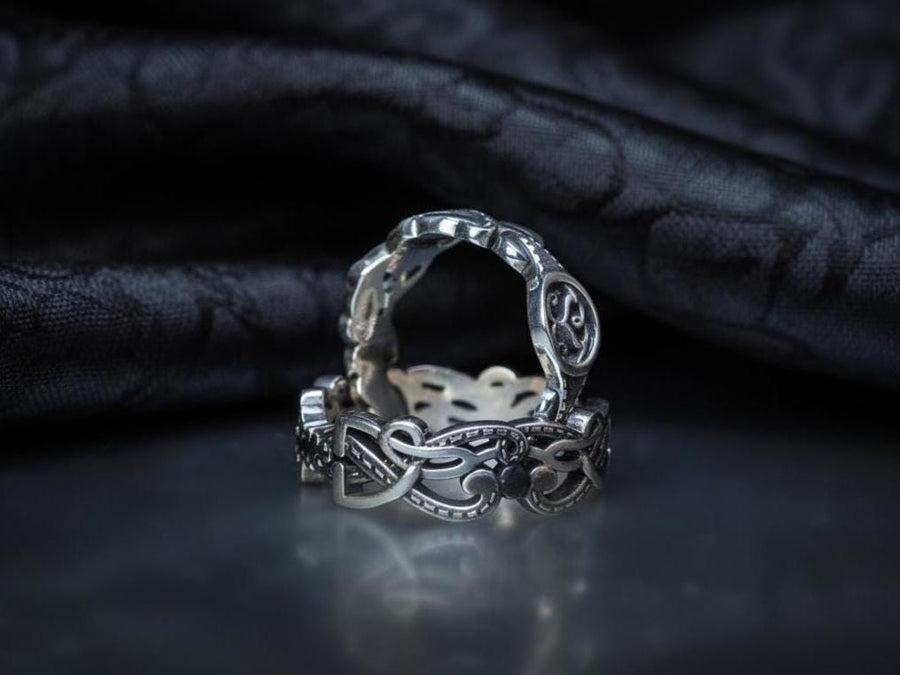 A studio shot of a matching two-piece sterling silver ring set on a black reflective surface. The standing ring is a signet style featuring an oval face with a raised Triskelion (triple spiral) motif. The ring lying flat is a wide band with an "openwork" or cut-out Celtic knot pattern. The silver is highly polished on the raised surfaces, contrasting with the dark shadows of the open areas.