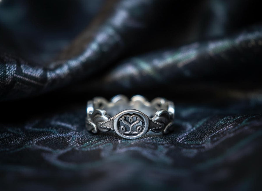 A cinematic macro shot of a sterling silver ring bathed in warm, golden light. The focal point is the central oval face, which encloses a raised relief design of two spirals meeting in the center to suggest a heart shape. The band shoulders feature textured knotwork that leads up to the centerpiece. The ring rests on a wet, reflective surface, creating a mirror image of the love knot below. (etsy image id: 7490345549)
