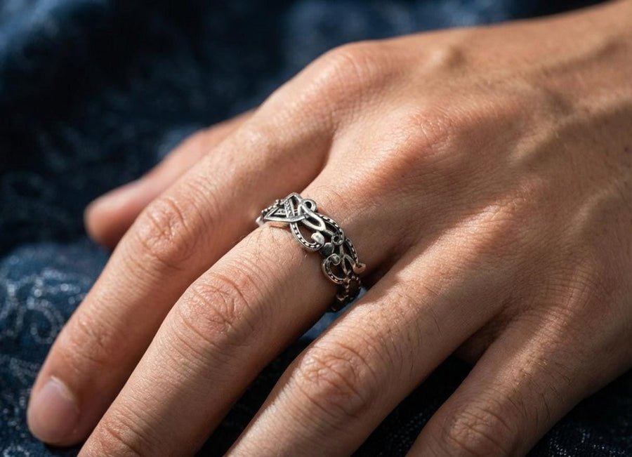 A cinematic close-up of a sterling silver ring lying on a reflective surface. The band features an "openwork" design, meaning the Celtic knot pattern is cut out, allowing light to pass through the loops. The silver strands weave over and under in a continuous infinity motif. The background is dark, but the ring is illuminated by a warm, golden mist. (etsy image id: 7442429936)