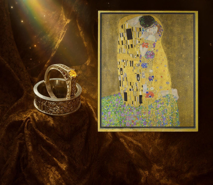 A cinematic studio shot of a matching two-piece ring set. The bottom ring is a wide silver band featuring a detailed relief carving of the couple from Gustav Klimt's "The Kiss." The top ring is a narrower engagement band that mimics the geometric patterns of the painting and holds a round, amber-colored gemstone. The background is a warm, golden haze, reflecting the "Golden Phase" of the original artwork.