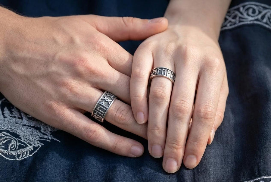 A clean studio shot of two matching sterling silver rings. The bands feature the 24 characters of the Elder Futhark runic alphabet wrapping around the exterior. The runes are polished silver, standing out sharply against a rough, blackened background texture. The rims are smooth and polished, framing the ancient script.