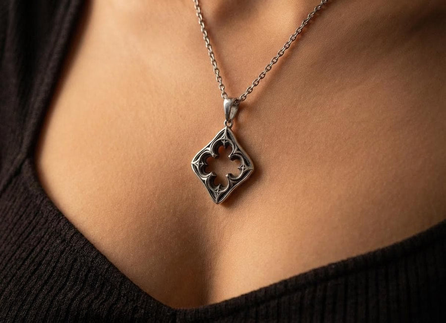 A cinematic studio shot of a diamond-shaped sterling silver pendant hanging against a smoky, amber-lit background. The design is "openwork," featuring a central cutout in the shape of a four-lobed quatrefoil. The silver frame mimics the stone mullions of a Gothic window, with pointed arches and sharp cusps. The metal is polished on the ridges and darkened on the sides to define the shape against the void. (etsy image id: 7496323581)