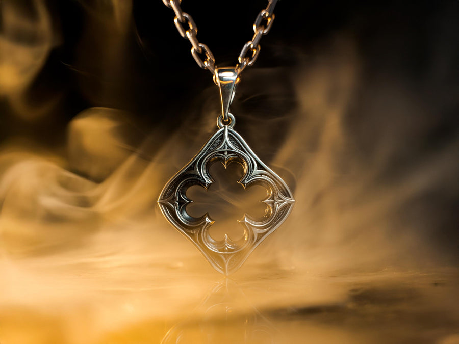 A cinematic studio shot of a diamond-shaped sterling silver pendant hanging against a smoky, amber-lit background. The design is "openwork," featuring a central cutout in the shape of a four-lobed quatrefoil. The silver frame mimics the stone mullions of a Gothic window, with pointed arches and sharp cusps. The metal is polished on the ridges and darkened on the sides to define the shape against the void. (etsy image id: 7496323581)
