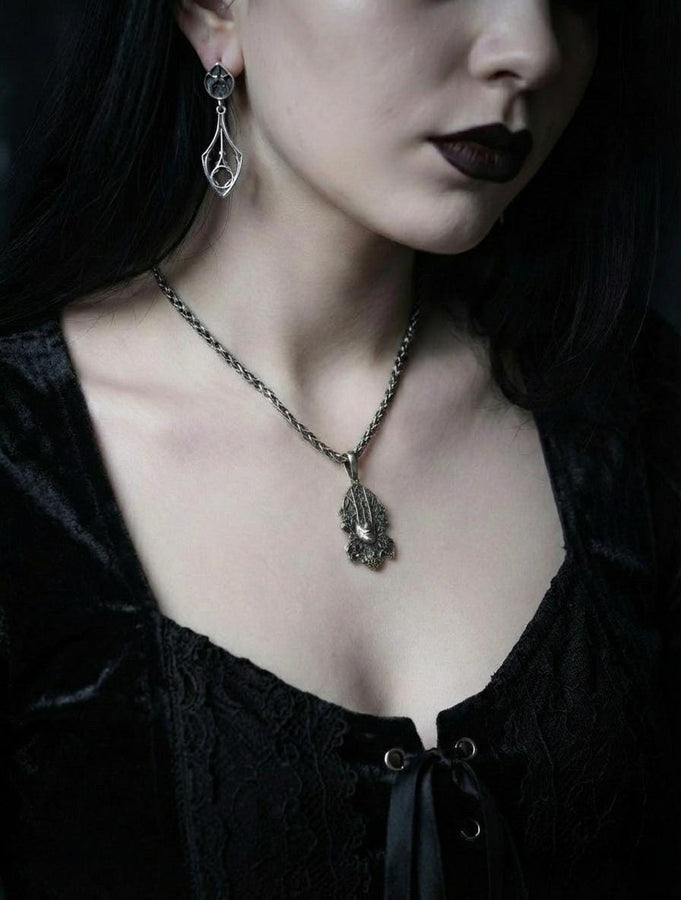 A detailed studio shot of a sterling silver pendant resting on a dark, textured stone surface. The design features a realistic human face in the center, but the forehead and eyes are obscured by a grid of Gothic window tracery (vertical bars and trefoils). The chin is framed by swirling Art Nouveau-style acanthus leaves. The silver is heavily oxidized, giving the face the appearance of a weathered stone statue. (etsy image id: 7448678222)