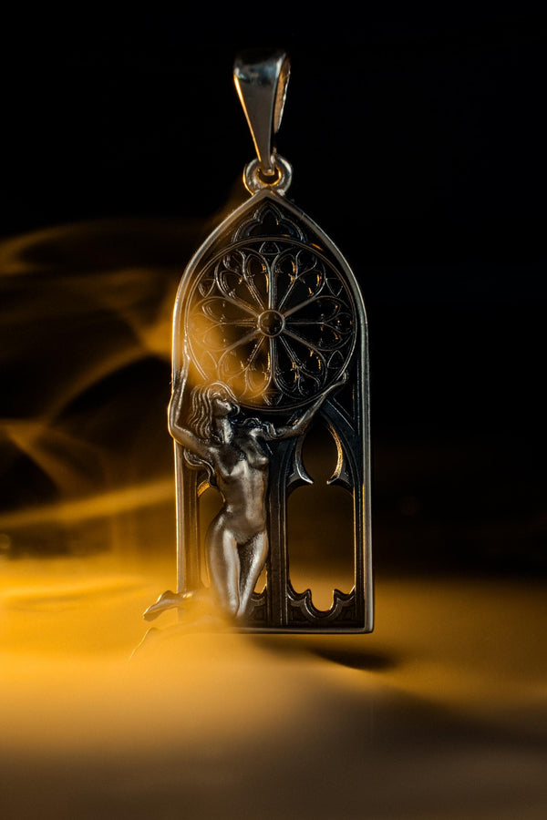A cinematic studio shot of a tall, arched sterling silver pendant. The frame mimics a Gothic cathedral window. Inside the arch, a sculpted female figure (appearing nude) kneels or floats, reaching up to hold a large, circular "Rose Window" tracery wheel above her head. The silver is heavily oxidized, creating deep shadows behind the figure and the window spokes. (etsy image id: 7448666740)