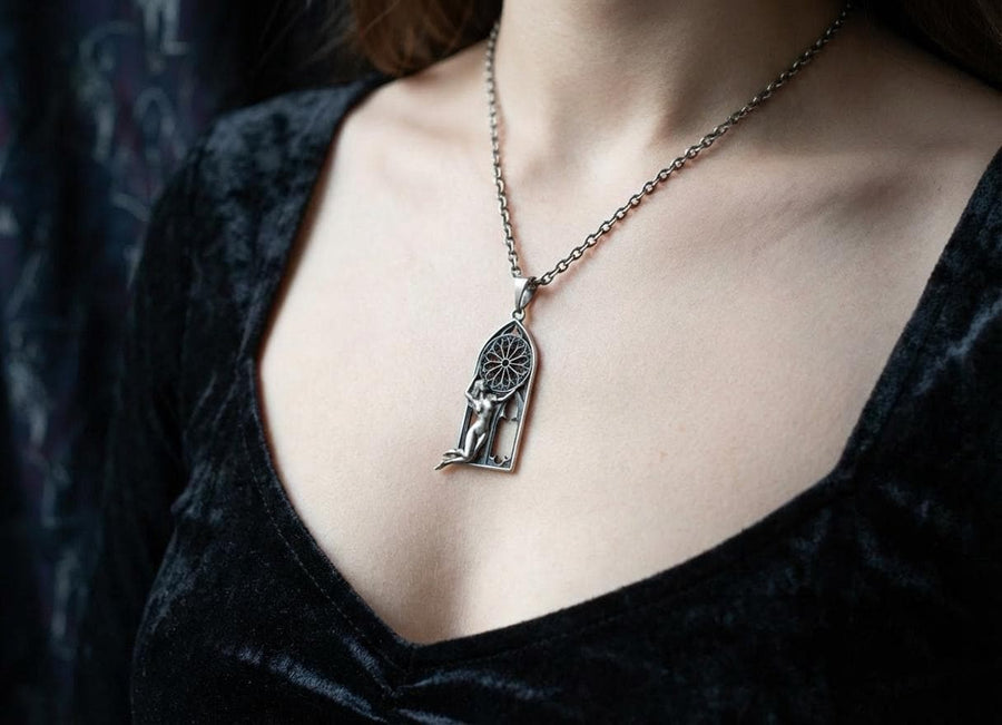 A cinematic studio shot of a tall, arched sterling silver pendant. The frame mimics a Gothic cathedral window. Inside the arch, a sculpted female figure (appearing nude) kneels or floats, reaching up to hold a large, circular "Rose Window" tracery wheel above her head. The silver is heavily oxidized, creating deep shadows behind the figure and the window spokes. (etsy image id: 7448666740)