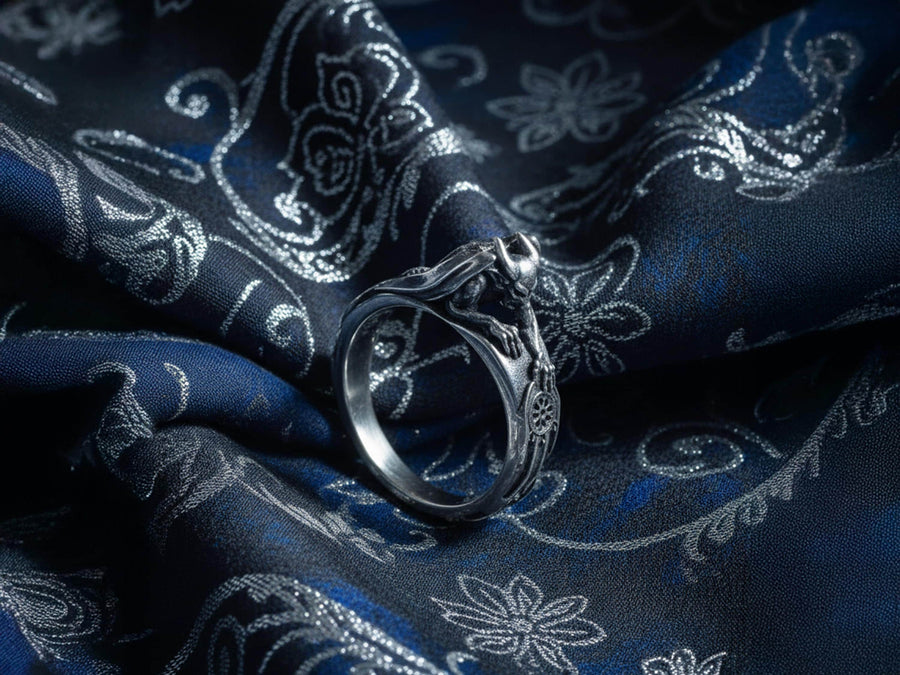 A detailed profile shot of a sculptural sterling silver ring. The band is formed by the body of a crouching gargoyle. The creature features distinct horns, a snarling face, and muscular limbs. Its bat-like wings fold back to form the sides of the ring shank. In its front claw, it holds a small, circular charm with a web pattern, resembling a dreamcatcher. The silver is heavily oxidized black in the crevices to accentuate the details. (etsy image id: 7497960785)