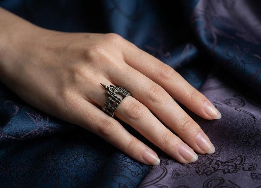 A cinematic studio shot of a sculptural sterling silver ring standing on a reflective surface. The ring is shaped like a miniature building, featuring a tall central spire that rises to a sharp point. The band itself is formed by a colonnade of pointed Gothic arches and buttresses that wrap around the finger. The silver is heavily oxidized, creating deep shadows in the "windows" and "doorways" of the structure. (etsy image id: 7450125356)