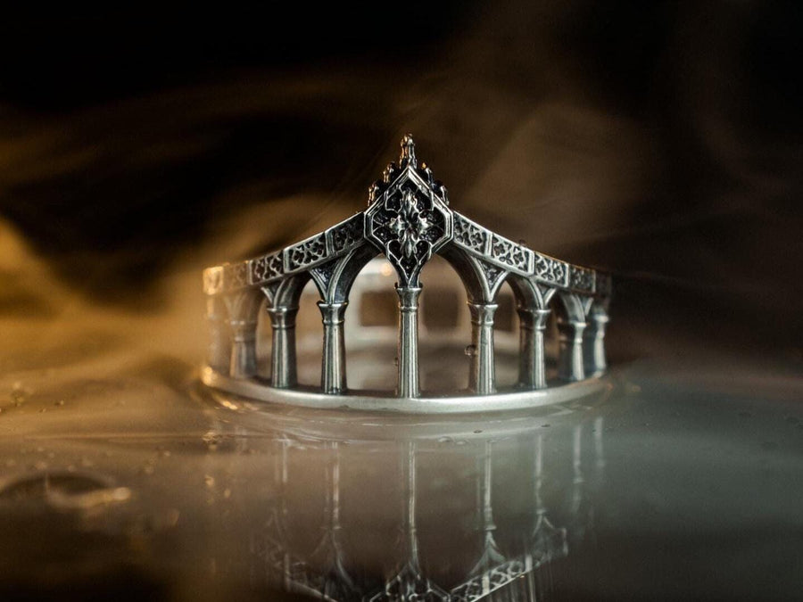 A cinematic studio shot of a sculptural sterling silver ring standing in shallow water. The design mimics a bridge or aqueduct, featuring a row of vertical pillars or columns that support a rising upper rail. The center rises to a sharp point adorned with a floral or quatrefoil relief. The space between the columns is open, creating a "see-through" architectural silhouette. (etsy image id: 7450247674)