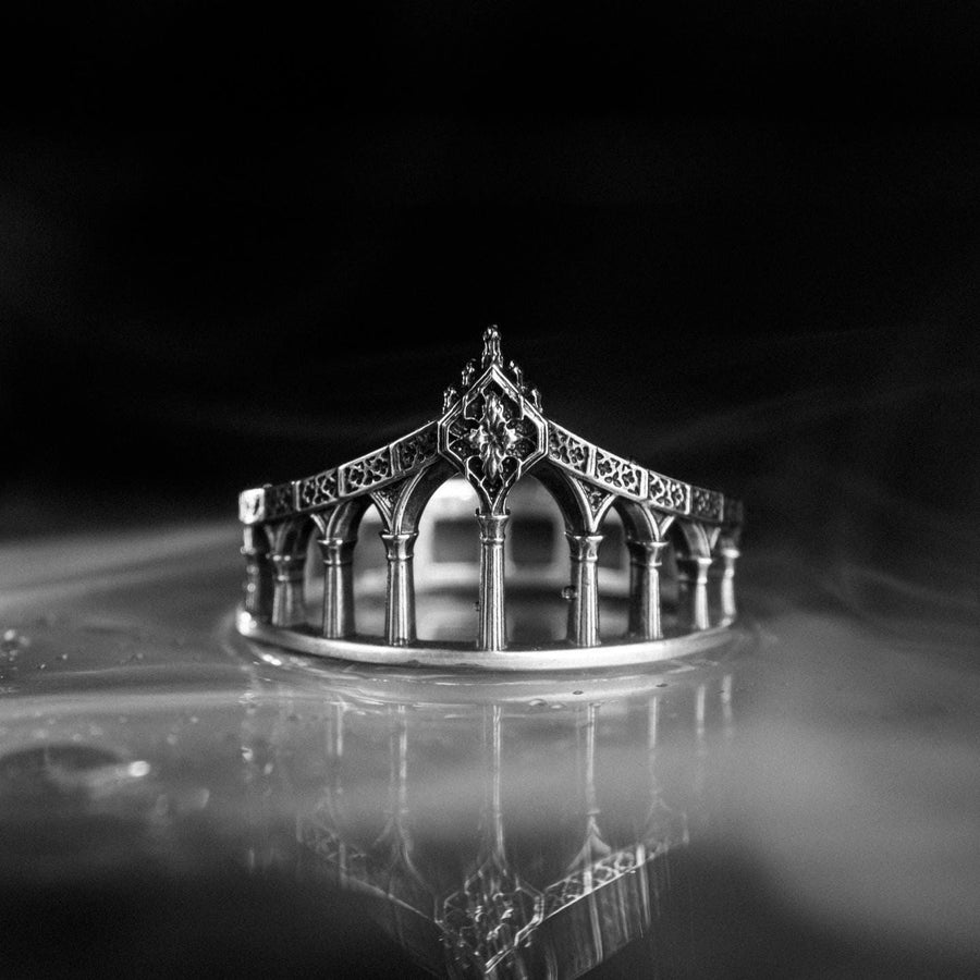 A cinematic studio shot of a sculptural sterling silver ring standing in shallow water. The design mimics a bridge or aqueduct, featuring a row of vertical pillars or columns that support a rising upper rail. The center rises to a sharp point adorned with a floral or quatrefoil relief. The space between the columns is open, creating a "see-through" architectural silhouette. (etsy image id: 7450247674)