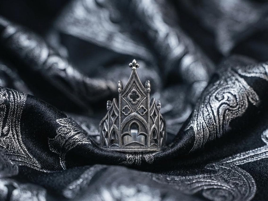 A clean studio shot of a sculptural sterling silver ring resting on a matte grey surface. The ring depicts a Gothic building facade with a tall, triangular central peak (gable) filled with geometric cross-shaped tracery. Flanking the center are lower pointed arches and vertical buttresses that wrap around the band. The silver is matte-finished with dark oxidation in the recesses to mimic stone shadows. (etsy image id: 7452370200)