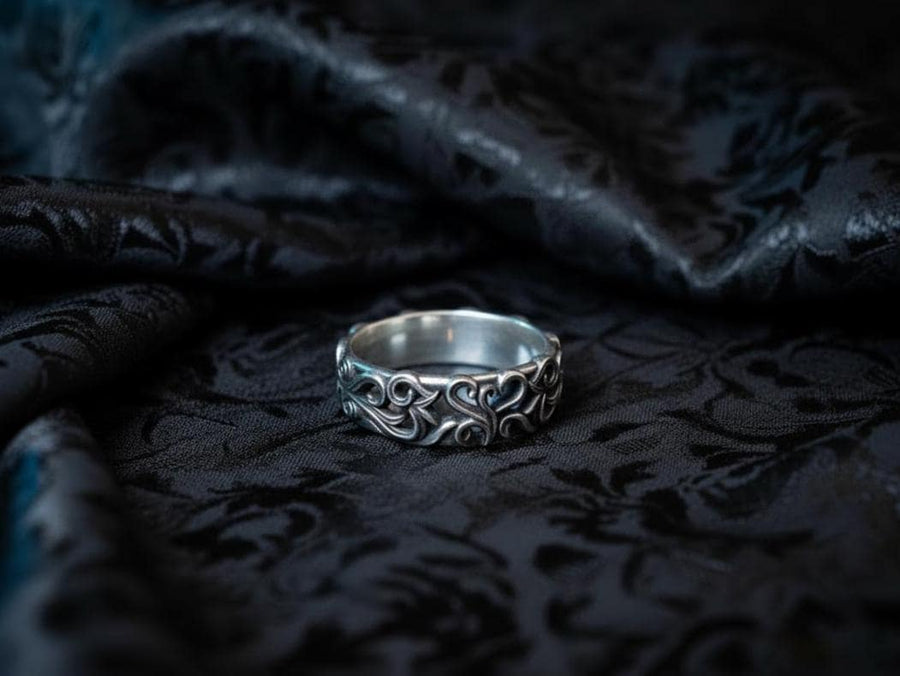 A detailed close-up of a wide sterling silver ring resting on a reflective surface. The band is covered in a deep-relief filigree pattern consisting of interlocking scrolls, vines, and subtle heart shapes. The raised designs are polished bright silver, while the recessed background is heavily oxidized to black, highlighting the complexity of the Baroque motif.