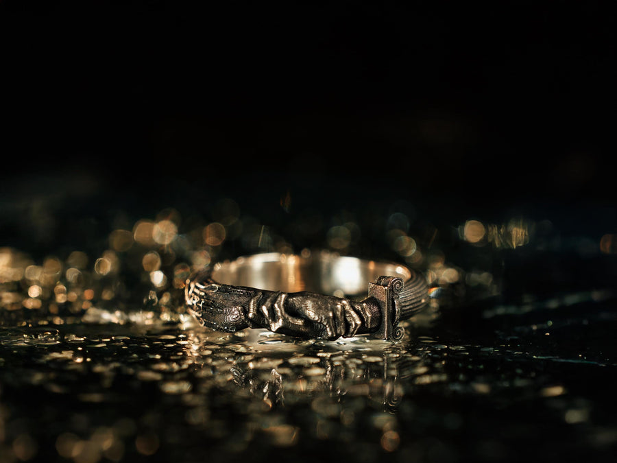 A cinematic studio shot of a sculptural sterling silver ring resting on a dark textured fabric. The focal point is two realistic hands clasped in a handshake (the Fede motif). Instead of fabric sleeves, the wrists emerge from architectural column capitals with distinct "volute" scrolls (Ionic style). The band continues as a textured, fluted column shaft. The silver is heavily oxidized to mimic ancient stone. (etsy image id: 7452415972)