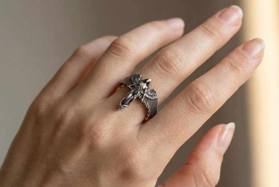 A clear studio shot of a sculptural sterling silver ring on a white background. The ring features a full-body female figure, likely an angel or goddess, positioned at the center. Her large, detailed wings spread horizontally to form the top half of the ring band. Her dress flows downward, tapering into the bottom of the band. (etsy image id: 7500519831)