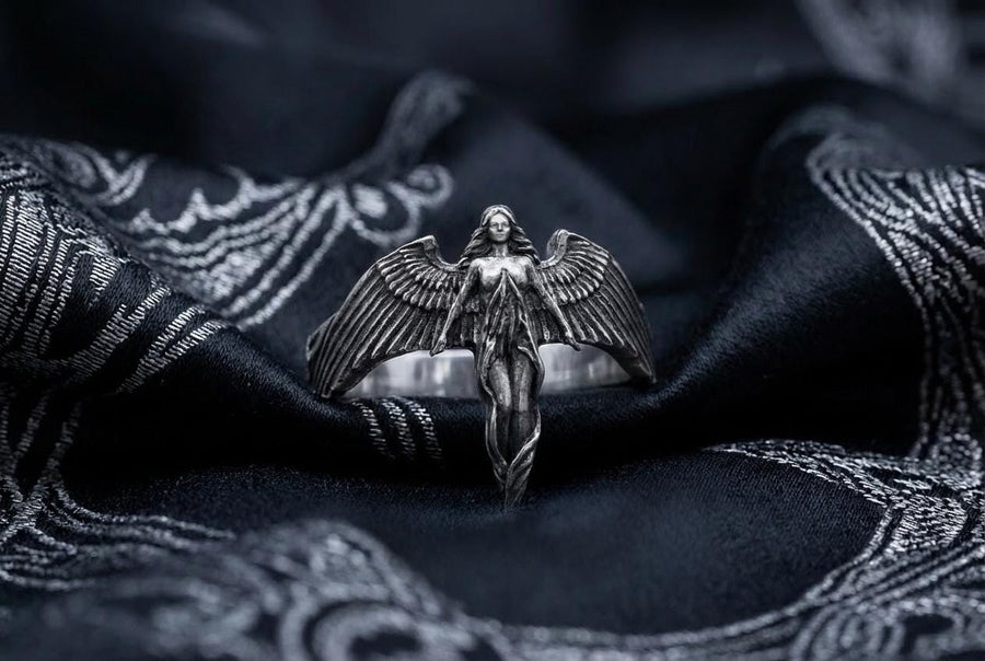 A clear studio shot of a sculptural sterling silver ring on a white background. The ring features a full-body female figure, likely an angel or goddess, positioned at the center. Her large, detailed wings spread horizontally to form the top half of the ring band. Her dress flows downward, tapering into the bottom of the band. (etsy image id: 7500519831)