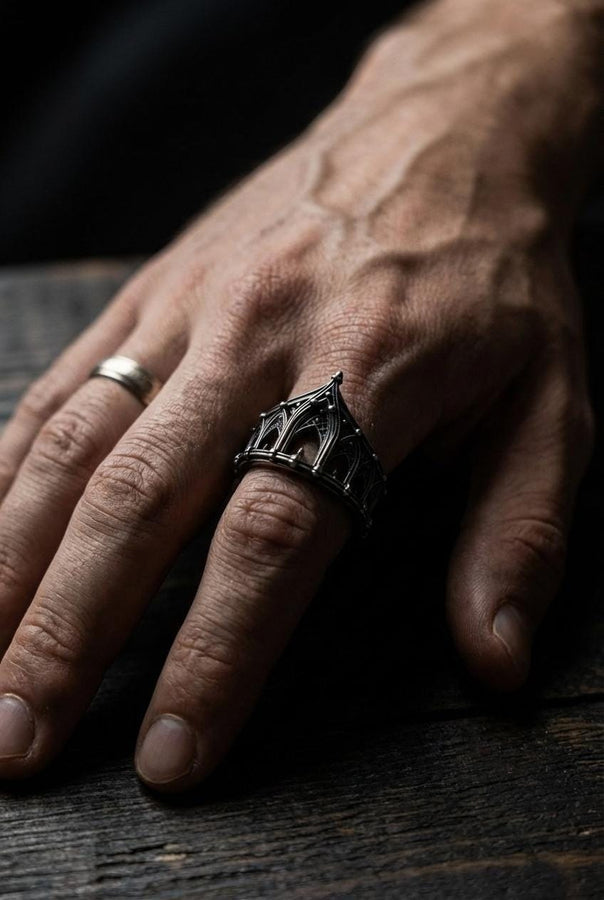 A cinematic studio shot of a sculptural sterling silver ring standing on a reflective surface. The ring is designed as a "crown" or "coronet," formed by a continuous circle of Gothic cathedral arches. The central arch is the tallest, creating a focal point like a spire. The silver is heavily oxidized, with black recesses emphasizing the ribbed vaulting and architectural details against the polished ridges.