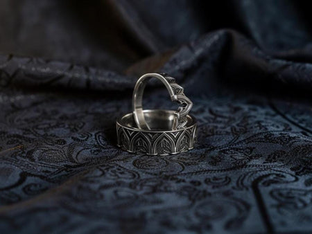 A cinematic studio shot of two matching sterling silver rings. The top ring is a delicate "crown" or tiara shape, formed by the peak of a Gothic arch with trefoil tracery. The bottom ring is a wide band featuring a continuous row of identical arches. The background metal inside the arches has a rough, "eroded" or stippled texture, heavily oxidized to black, while the raised architectural lines are polished smooth.