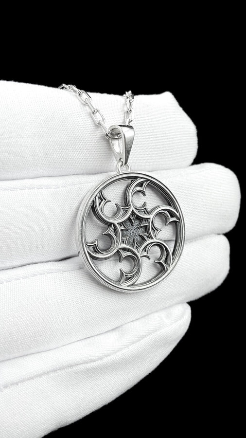 A detailed close-up of a circular sterling silver pendant resting on a dark blue, textured silk fabric. The pendant features an openwork design with swirling, crescent-like shapes radiating from a central four-pointed star. The intricate pattern resembles a gothic rose window or a celestial wheel. The raised metal surfaces are polished to a bright silver shine, contrasting beautifully against the dark, oxidized background recesses. (etsy image id: 7830974908)