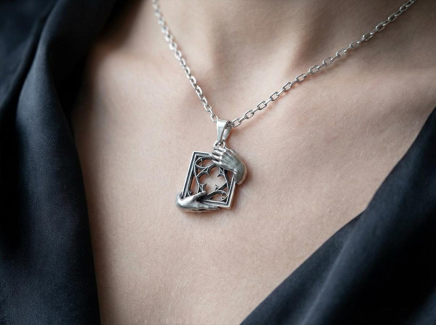 A close-up studio photograph of a highly detailed sterling silver pendant resting on a luxurious, dark blue silk fabric with silvery thread patterns. The pendant's striking design features two realistically sculpted, three-dimensional hands emerging from the sides to gently grasp a rectangular Gothic window frame. Inside the frame sits intricate openwork tracery, forming a classic quatrefoil motif. The polished silver highlights contrast with (etsy image id: 7879251833)