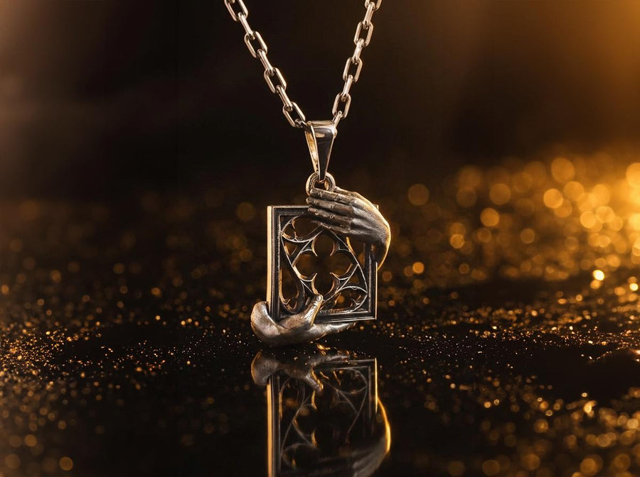 A close-up studio photograph of a highly detailed sterling silver pendant resting on a luxurious, dark blue silk fabric with silvery thread patterns. The pendant's striking design features two realistically sculpted, three-dimensional hands emerging from the sides to gently grasp a rectangular Gothic window frame. Inside the frame sits intricate openwork tracery, forming a classic quatrefoil motif. The polished silver highlights contrast with (etsy image id: 7879251833)