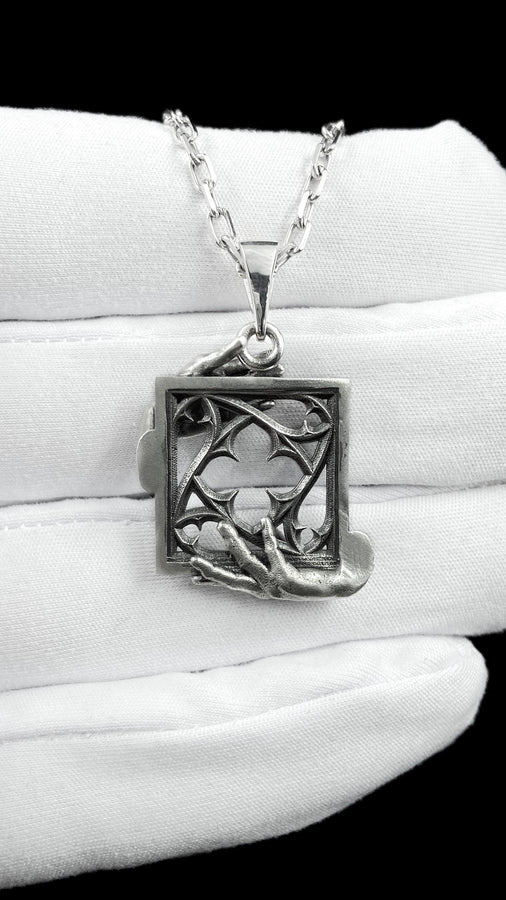 A close-up studio photograph of a highly detailed sterling silver pendant resting on a luxurious, dark blue silk fabric with silvery thread patterns. The pendant's striking design features two realistically sculpted, three-dimensional hands emerging from the sides to gently grasp a rectangular Gothic window frame. Inside the frame sits intricate openwork tracery, forming a classic quatrefoil motif. The polished silver highlights contrast with (etsy image id: 7879251833)