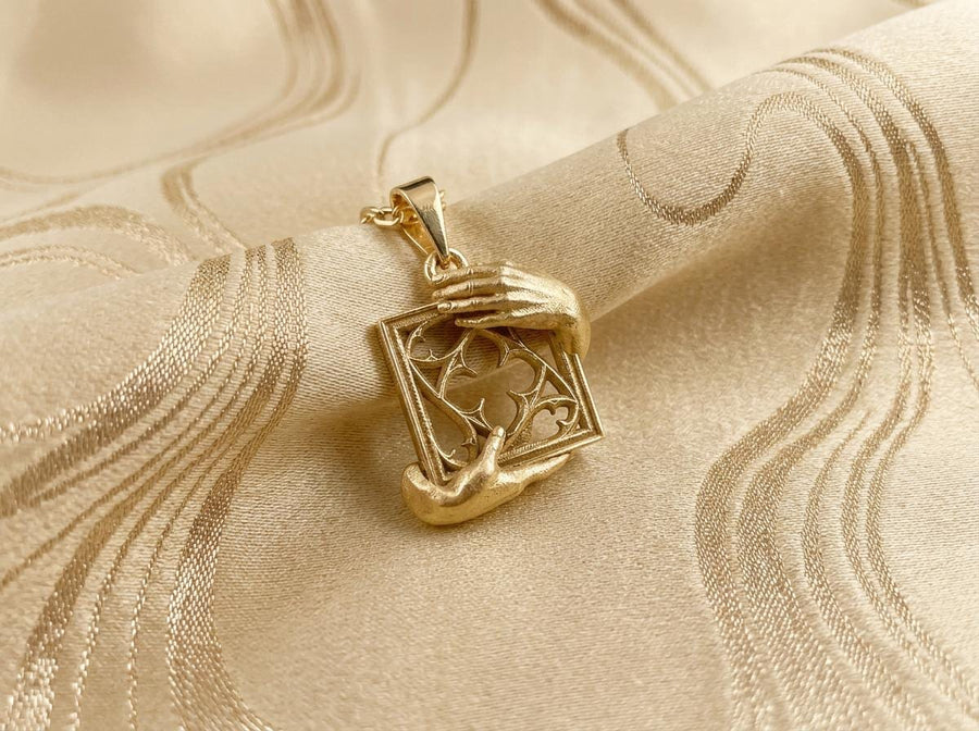 A close-up studio photograph of a highly detailed sterling silver pendant resting on a luxurious, dark blue silk fabric with silvery thread patterns. The pendant's striking design features two realistically sculpted, three-dimensional hands emerging from the sides to gently grasp a rectangular Gothic window frame. Inside the frame sits intricate openwork tracery, forming a classic quatrefoil motif. The polished silver highlights contrast with (etsy image id: 7879251833)