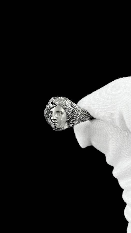 A high-contrast macro photograph of a heavy sterling silver ring resting against a pitch-black background. The ring features a three-dimensional sculpture of a classical Greek goddess's face, likely Athena. A jagged, deep crack splits the face vertically. The right side shows smooth, classical features, while the left side transitions into a rough, eroded, and porous texture resembling crumbling ancient stone. The silver is heavily oxidized t (etsy image id: 7831406732)