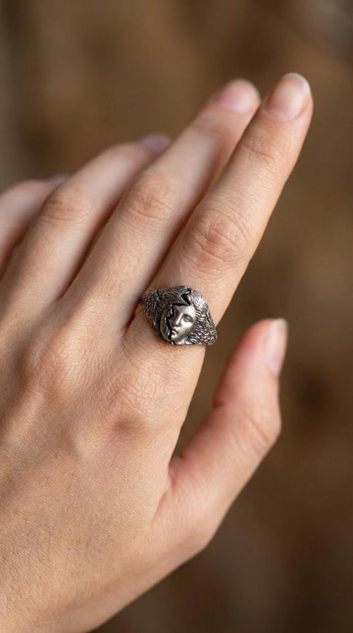 A high-contrast macro photograph of a heavy sterling silver ring resting against a pitch-black background. The ring features a three-dimensional sculpture of a classical Greek goddess's face, likely Athena. A jagged, deep crack splits the face vertically. The right side shows smooth, classical features, while the left side transitions into a rough, eroded, and porous texture resembling crumbling ancient stone. The silver is heavily oxidized t (etsy image id: 7831406732)